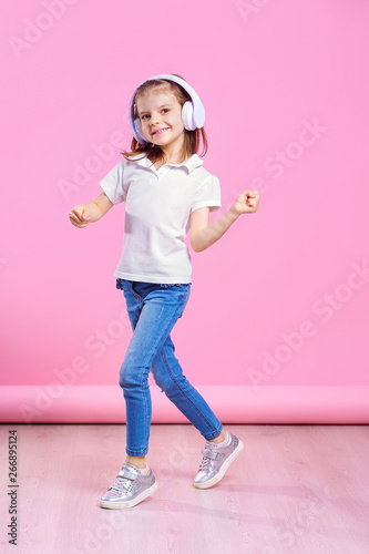 Girl listening to music in headphones an dance. Cute child enjoying happy dance music, smile, posing on pink studio background wall. © zamuruev