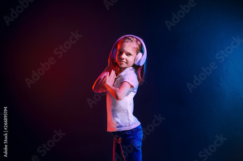 Girl of 7 years old listening to music in headphones on dark colorful background. Neon light. Cute child enjoying happy dance music.