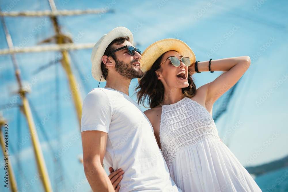 Happy young couple walking by the harbor of a touristic sea resort with sailboats on background