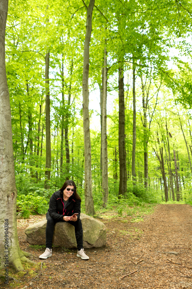 Lady sits on a cut tree trunk in nature while interacting with her smartphone Lady sits on a cut tree trunk in nature while interacting with her smartphone