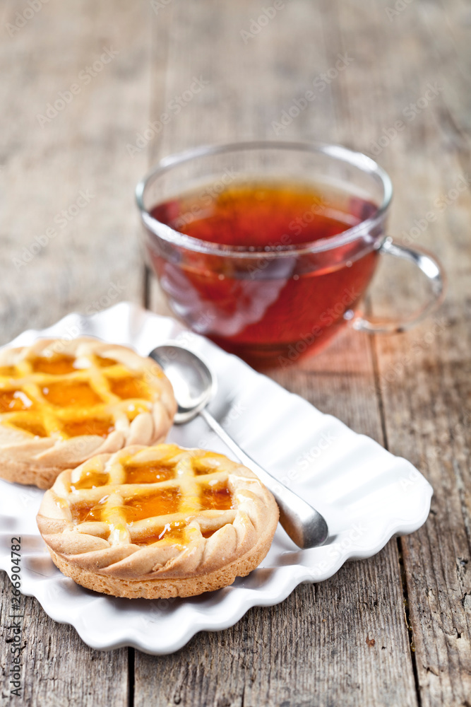 Fresh baked tarts with marmalade or apricot jam filling on white ceramic plate and cup of tea on rustic wooden table. Fresh baked tarts with marmalade or apricot jam filling on white ceramic plate and cup of tea on rustic wooden table.