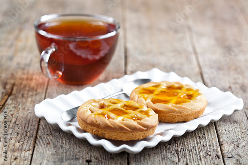 Fresh baked tarts with marmalade or apricot jam filling on white ceramic plate and cup of tea on rustic wooden table. photo