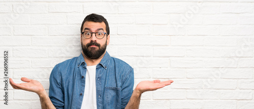 Handsome man with beard over white brick wall having doubts while raising hands photo