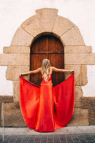 The girl in the red. A slender girl in a tight long red fluttering dress with bare shoulders. Standing in the street near the big old wooden door framed with stone. 