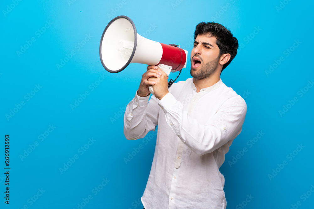 Young man over isolated blue wall shouting through a megaphone Young man over isolated blue wall shouting through a megaphone