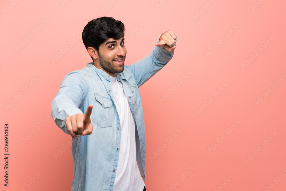 Young man over pink wall points finger at you while smiling Young man over pink wall points finger at you while smiling
