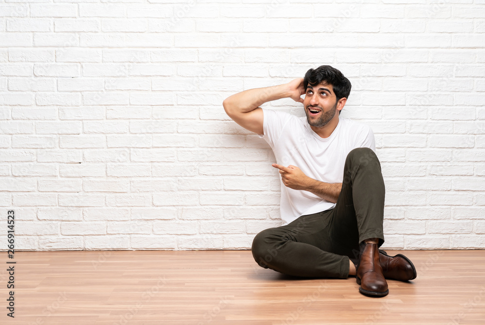 Young man sitting on the floor surprised and pointing finger to the side Young man sitting on the floor surprised and pointing finger to the side