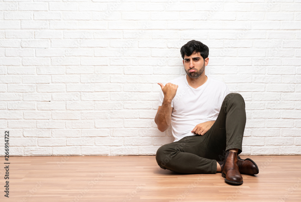 Young man sitting on the floor unhappy and pointing to the side