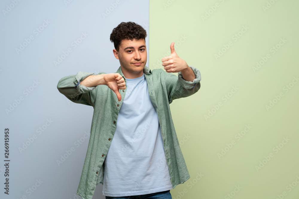 Young man over blue and green background making good-bad sign. Undecided between yes or not Young man over blue and green background making good-bad sign. Undecided between yes or not