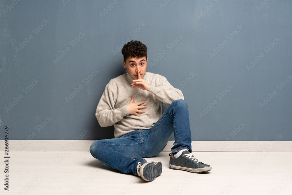 Young man sitting on the floor showing a sign of silence gesture putting finger in mouth Young man sitting on the floor showing a sign of silence gesture putting finger in mouth