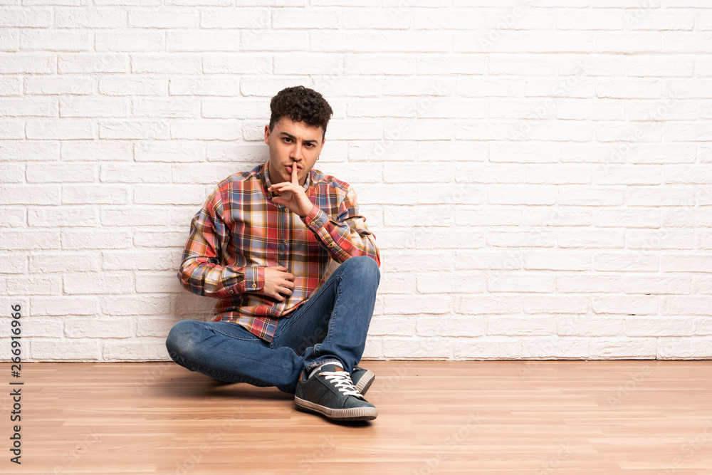 Young man sitting on the floor showing a sign of silence gesture putting finger in mouth Young man sitting on the floor showing a sign of silence gesture putting finger in mouth
