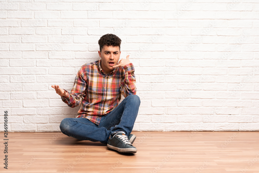 Young man sitting on the floor making phone gesture and doubting