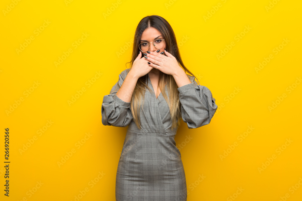 Young woman with glasses over yellow wall covering mouth with hands for saying something inappropriate Young woman with glasses over yellow wall covering mouth with hands for saying something inappropriate
