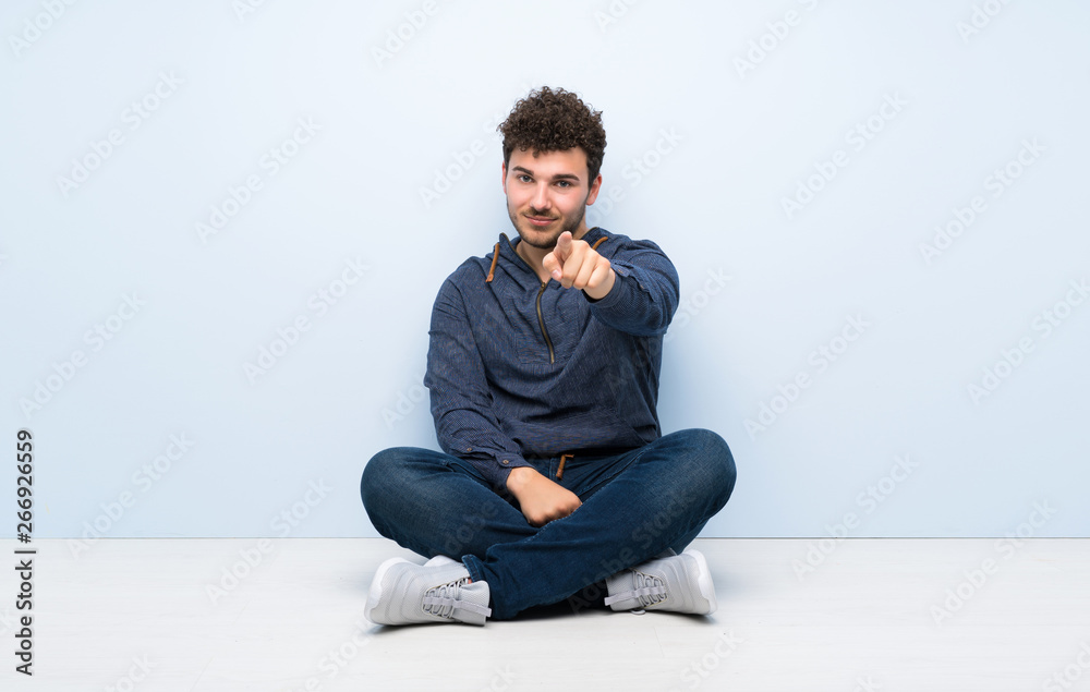 Young man sitting on the floor points finger at you with a confident expression Young man sitting on the floor points finger at you with a confident expression