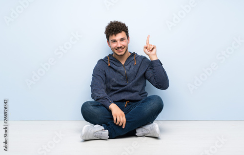Young man sitting on the floor showing and lifting a finger in sign of the best