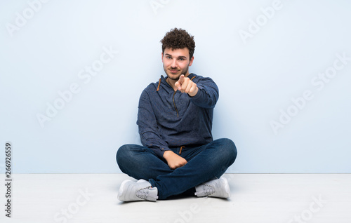 Young man sitting on the floor points finger at you with a confident expression © luismolinero