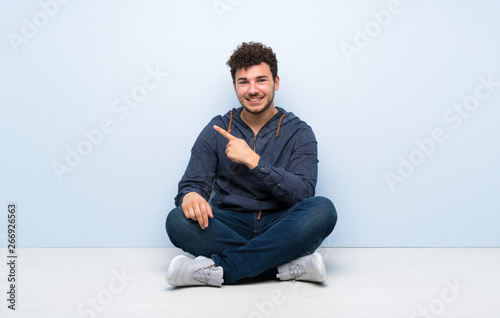 Young man sitting on the floor pointing to the side to present a product © luismolinero