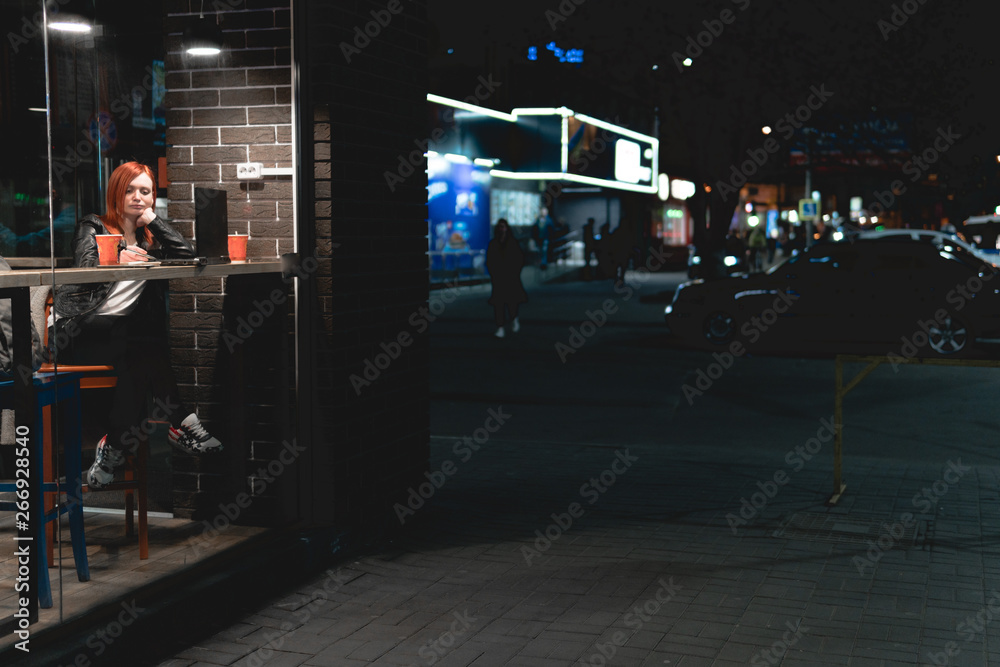 Businesswoman, girl working on laptop in cafe, hold smartphone in hands, pen, use phone. Freelancer works remotely. Online, network, education. Night city street, dark theme. tea cup, copy space