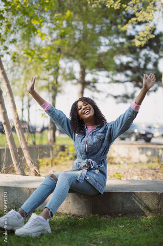 Happy african american woman sitting in a park with raised hands having fun