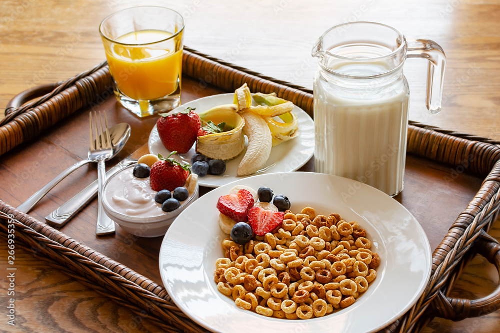 A bowl of oat cereal on wooden tray with assorted fruit, yogurt, orange juice and milk.  Tray on wooden table.  Angled side view.