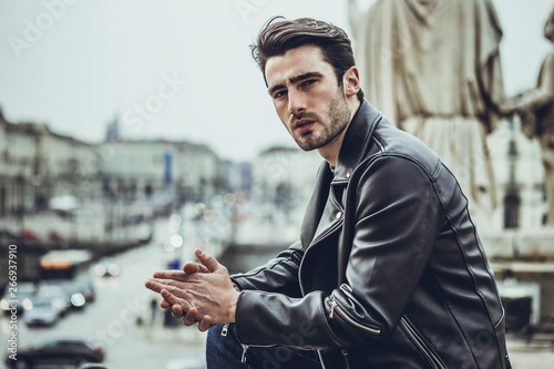One handsome young man in urban setting in modern city, standing, wearing black leather jacket and jeans, looking at camera