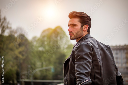 One handsome young man in urban setting in modern city, standing, wearing black leather jacket and jeans, looking away