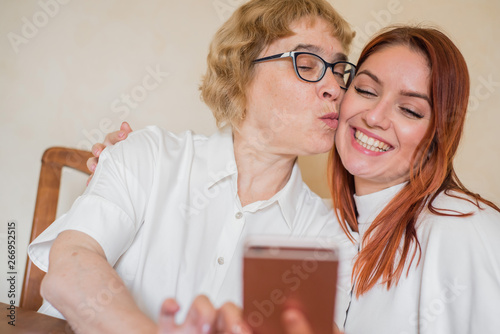 An elderly mother and daughter dressed in light clothes are smiling and taking selfies. Mom and daughter are photographed on the front camera of the smartphone while sitting at home at the table.