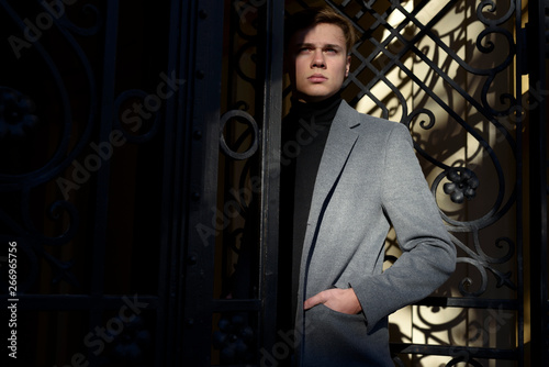 Stylish man in black polo neck and melange grey jacket, standing in decorative metal gate, a bit in sunlight and shadow.