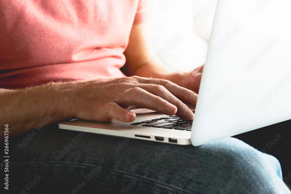 Fototapeta premium a man in jeans and a t-shirt sits on the couch and prints on a laptop. men's hands, print on the computer, close-up