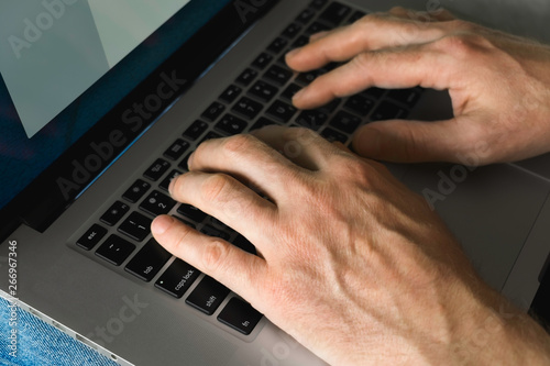 a man in jeans and a t-shirt sits on the couch and prints on a laptop. men's hands, print on the computer, close-up