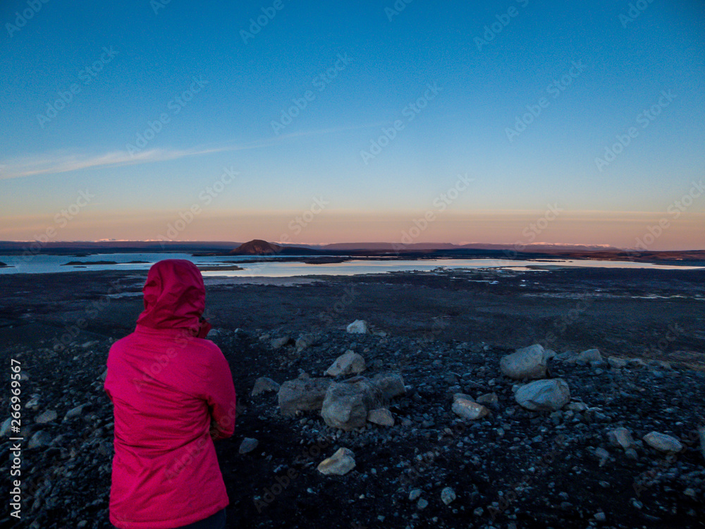 Girl wearing pink jacket walks around the top of the volcano. Sunrise on the horizon. Ground is covered with volcanic stones. In the back foam is coming out of the ground. Geothermal energy region.