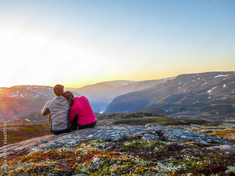 A couple sitting at the rock and watching the sunset. Sun sets behind the mountains. Sky is full of colors. Girl is leaning on the boy. Happiness, passion and love joined with the pursue of travel.