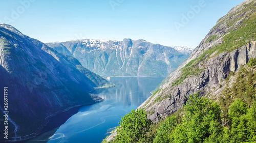 An majestic view on Eidfjord from Kjeasen, Norway. Slopes of the mountains are overgrown with lush green grass. Water has dark blue color. Taller parts of the mountains are barren. Sunny and clear day