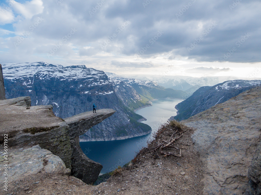 A man wearing blue jacket stands at the hanging rock formation, Trolltunga with a view on Ringedalsvatnet lake, Norway. Slopes of the mountains are partially covered with snow. Freedom and happiness