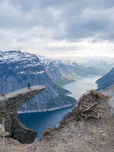 A man wearing blue jacket stands at the hanging rock formation, Trolltunga with a view on Ringedalsvatnet lake, Norway. Slopes of the mountains are partially covered with snow. Freedom and happiness photo