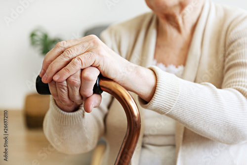 Elderly woman sitting in nursing home room holding walking quad cane with wrinked hand. Old age senior lady wearing beige cardigan, metal aid stick handle bar close up. Interior background, copy space