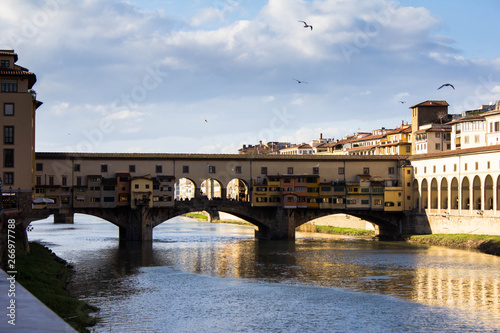 The Ponte Vecchio, medieval stone ancient bridge over the Arno river in Florence, Italy. Bridge with balconies and residential spaces. Blue sky above bridge, seagulls flying. Italian old architecture.