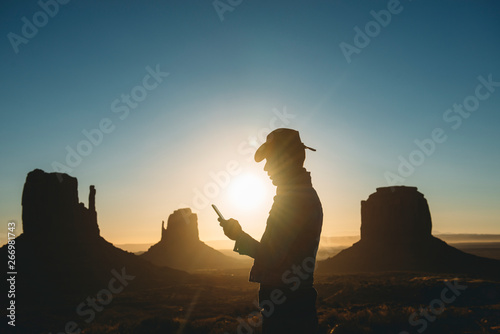 USA, Utah, Monument Valley, silhouette of man with cowboy hat looking at mobile phone at sunrise