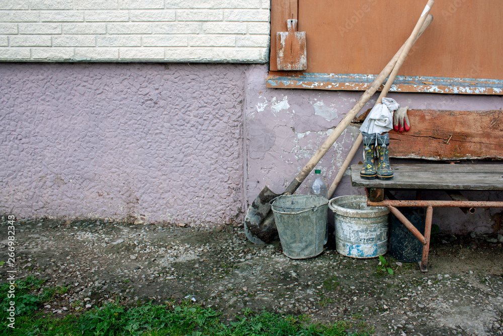 Two old buckets and shovels stand near a bench by the wall of the house. Spring work in the village. Black earth on a spade and green grass in a village in spring. Two old buckets and shovels stand near a bench by the wall of the house. Spring work in the village. Black earth on a spade and green grass in a village in spring.