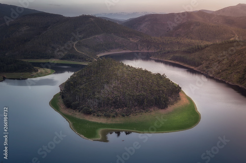 Aerial view of wooded island on smooth surface of lake in deserted undulating terrain