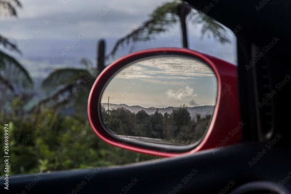 View in the wing mirror of a red sports racing car. Mirror is in focus and shows clouds over a bush scene from the Waitakere Ranges in Auckland. The background is blurred tree ferns or punga.
