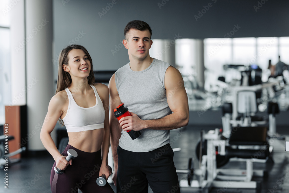 Attractive young sports people are holding bottle of water, talking and smiling while resting in gym Attractive young sports people are holding bottle of water, talking and smiling while resting in gym