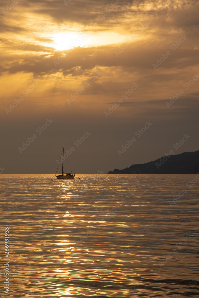 Backlight view of a sail boat anchored in the Bay of Fairy Tales at sunset with sun rays filtering through the clouds and the coastline in the background, Sestri Levante, Liguria, Italy