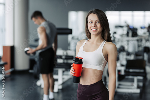 Attractive sport girl smiling and drinking water while standing at the gym with the boy training on background.