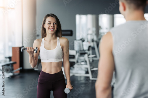 Young beautiful woman doing exercises with dumbbell in gym. Glad smiling girl is enjoying with her training process. She is working hard. Her trainer looking at this process.