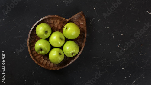Green apples in wooden carved bowl on black working board, photographed from above. © Lubo Ivanko