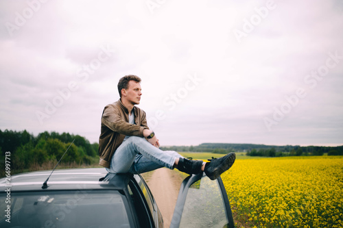 cool young man in a leather jacket is sitting on the roof of a car on the nature in a yellow field of flowers
