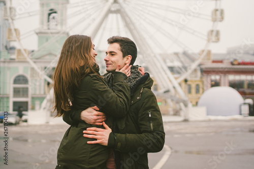 Theme love and holiday Valentines Day. pair of Caucasian heterosexual lovers in winter together gloomy weather embrace against background of Ferris wheel in town square. The guy gently hugs the girl