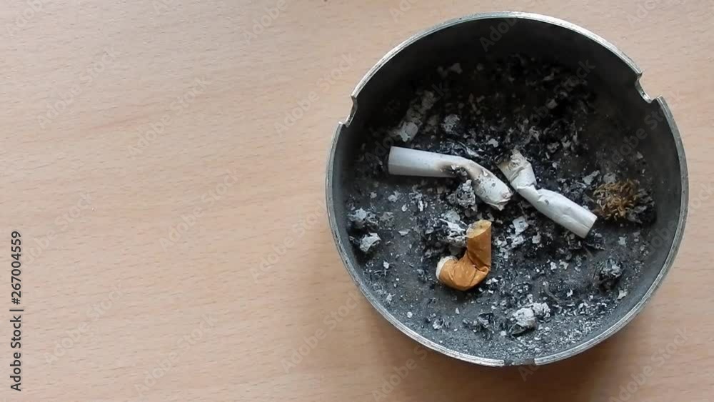 Close up over top of cigarette, smoking in ash tray. Hand takes & replaces cigarette to leave it smoking toxic fumes in ashtray with finished cigarettes. Room for copy to left.