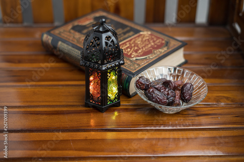 Islamic, ramadan and praying concept. Arabic lantern, dates or kurma with arabic book known as kitab or Quran on classic shine wood background, copy space, selective focus.